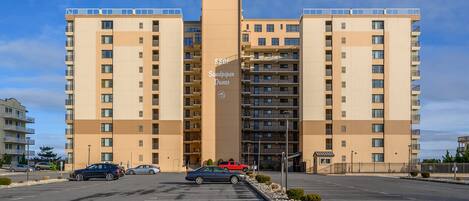 A multi-story residential building with a central entrance, parking lot, and a clear sky.