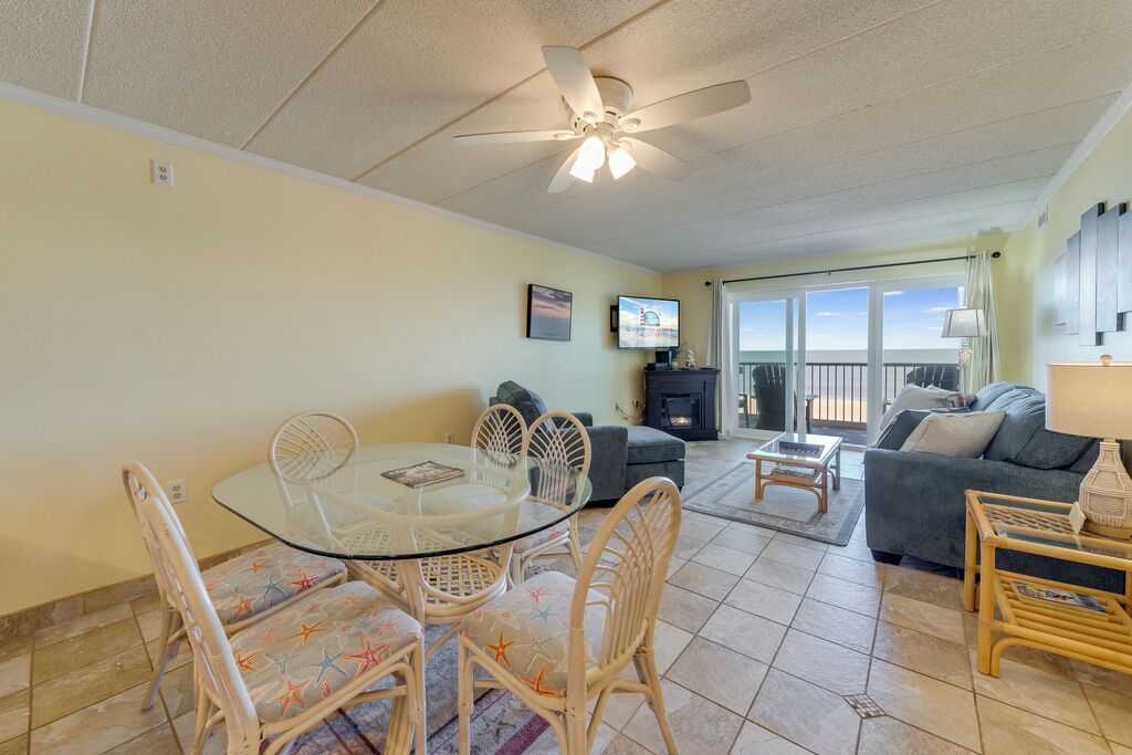 A spacious living room with a glass dining table, wicker chairs, a sofa, and a fireplace.