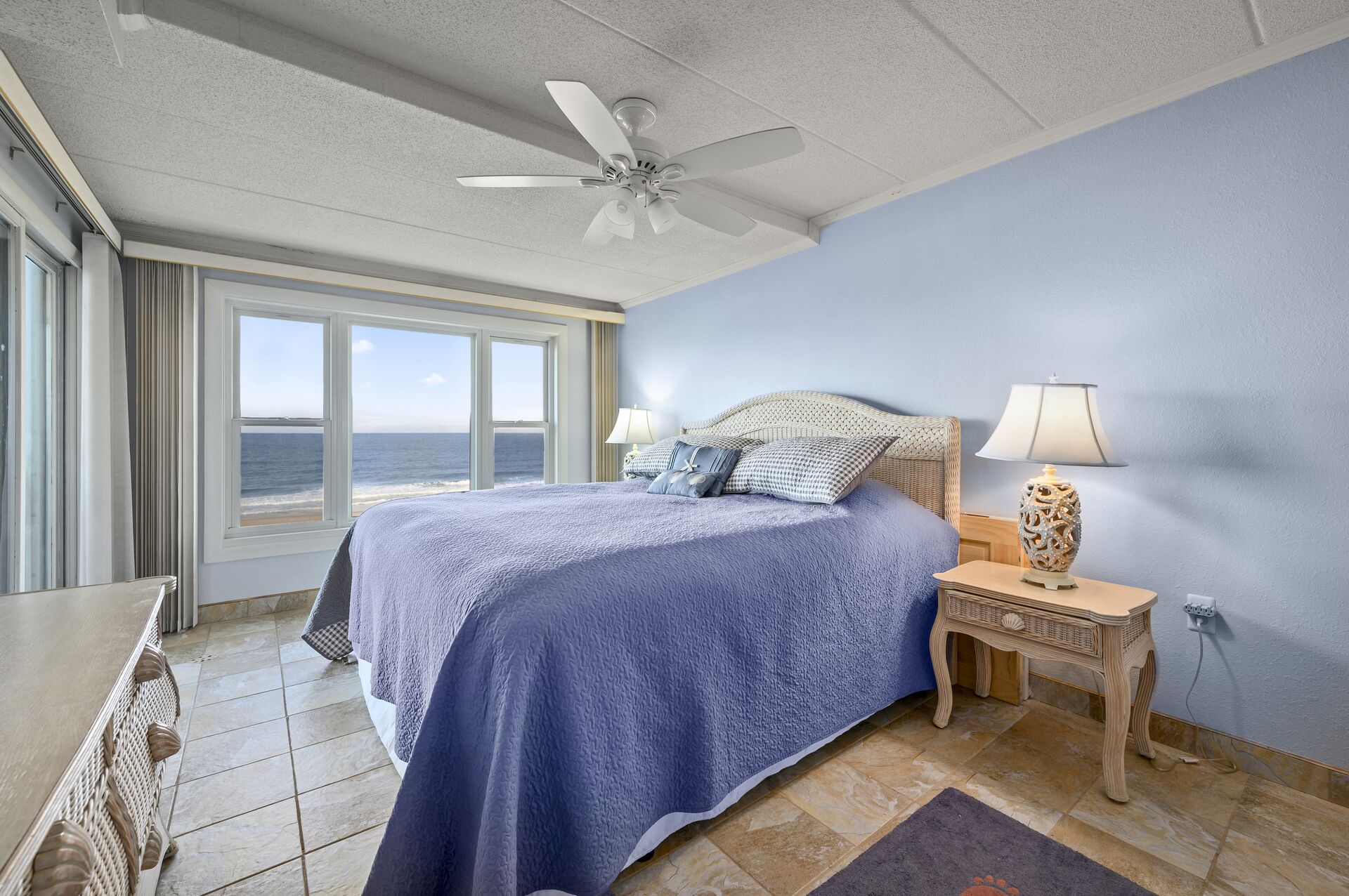 Une chambre à coucher avec un lit, une table de chevet, une lampe, un ventilateur de plafond et une vue sur l’océan.
