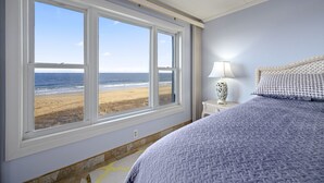 Une chambre avec une grande fenêtre donnant sur une plage, une lampe de chevet et un lit recouvert d’une couverture à motifs.