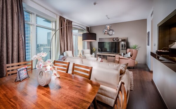 A modern living room with a wooden dining table, sofas, a TV, and a large window with curtains.