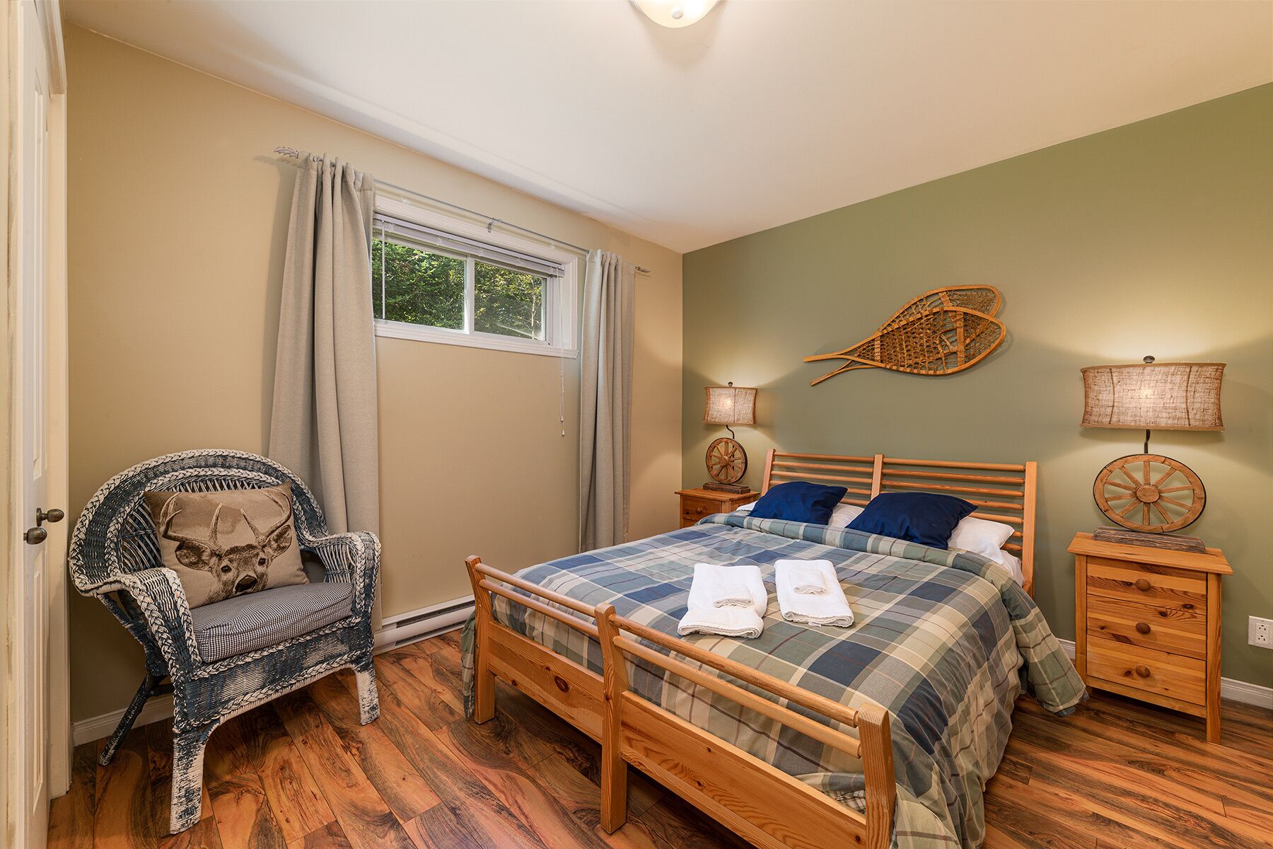 A bedroom with a wooden bed, a wicker chair, two bedside tables with lamps, and a window with curtains.
