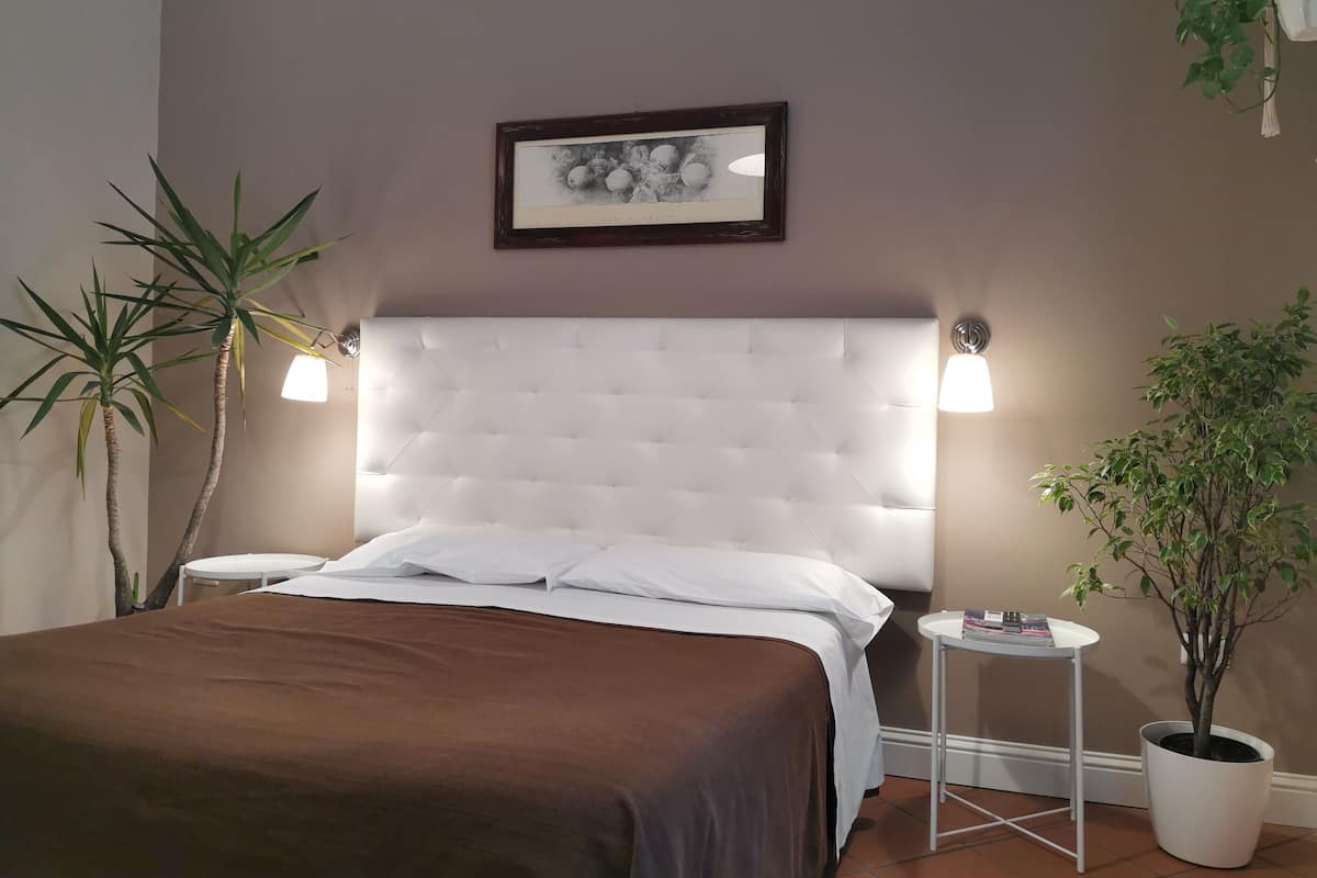 Una cama bien hecha con cabecera capitonada, dos mesitas de noche con lámparas, un cuadro enmarcado en la pared y plantas en macetas.