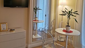 Ein modernes Hotelzimmer mit Flachbildfernseher, einem großen Spiegel, einem runden Esstisch mit Stühlen, einem kleinen Beistelltisch und einer Topfpflanze.