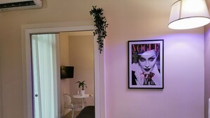 Ein Zimmer mit einem Bett, einem kleinen Tisch mit einer Pflanze, einem gerahmten Magazin an der Wand und einem an der Wand befestigten Fernseher.