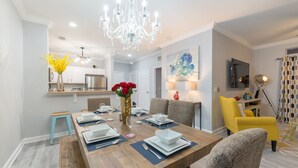 Un comedor con mesa de madera dispuesta para cuatro personas, rodeada de sillas tapizadas. Un candelabro cuelga del techo, y se aprecia una zona de cocina al fondo.