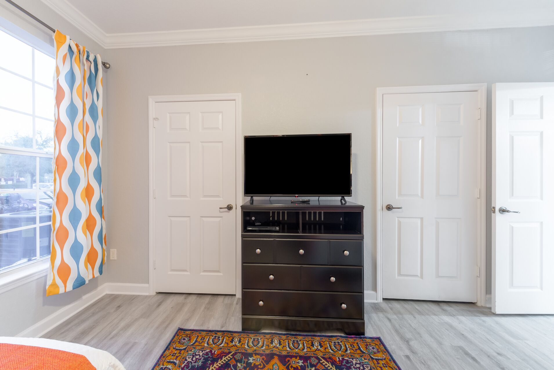 Un dormitorio con un televisor sobre una cómoda, dos puertas blancas y una alfombra estampada.