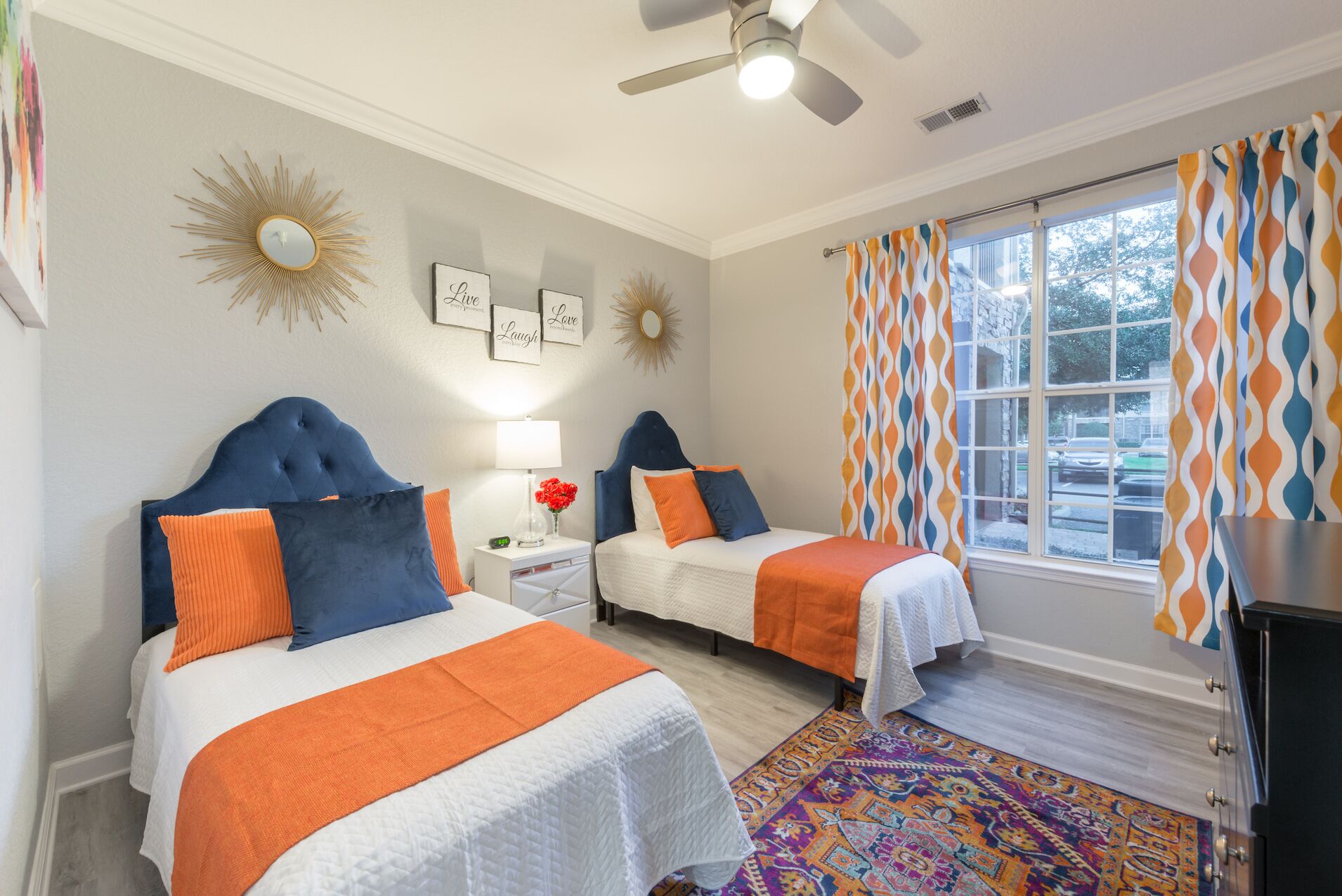 A hotel room with two beds, a ceiling fan, a window with patterned curtains, and a decorative wall piece.