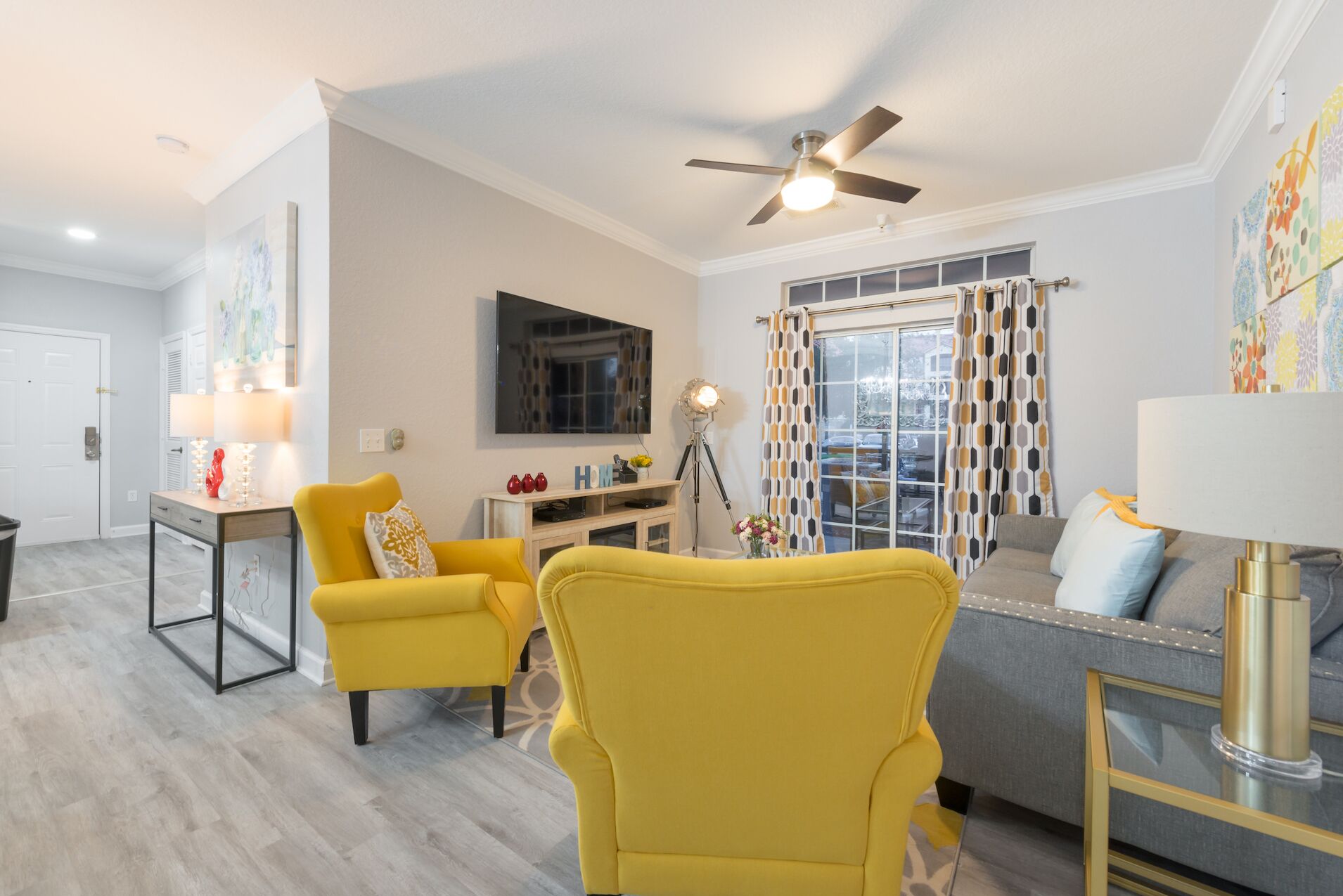 Un salon moderne avec un fauteuil jaune, un canapé gris, un ventilateur de plafond et une grande fenêtre ornée de rideaux à motifs.