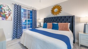 Un dormitorio con una cama grande, un ventilador de techo, una ventana con cortinas estampadas y un espejo montado en la pared.