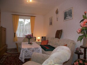 Ein gemütliches Wohnzimmer mit einer Couch, einem Tisch mit weißen Spitzen, einer Lampe und einer Vase mit Blumen.