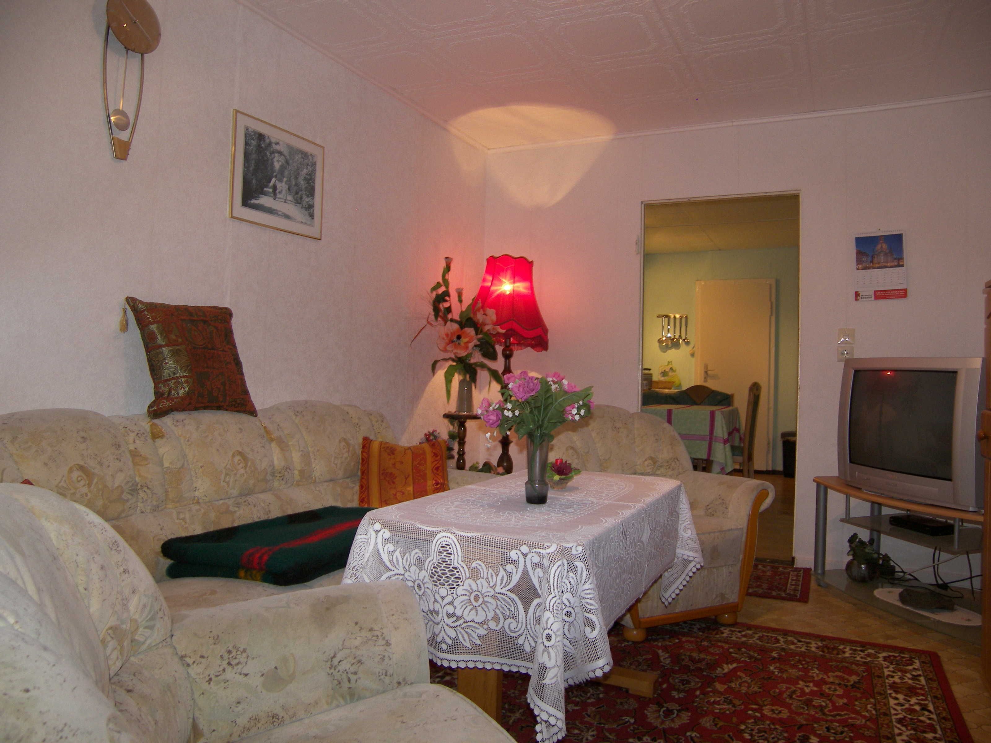 Ein Wohnzimmer mit einer Couch, einem Tisch mit weißer Spitze, einer Lampe und einem Fernseher.