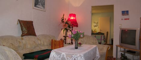 Uma sala de estar com um sofá, uma mesa com toalha rendada, um abajur e uma televisão.