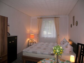 Ein Schlafzimmer mit einem Bett, einem Nachttisch mit Lampe, einer Kommode, einem Fenster mit Vorhängen und einer Vase mit Blumen.