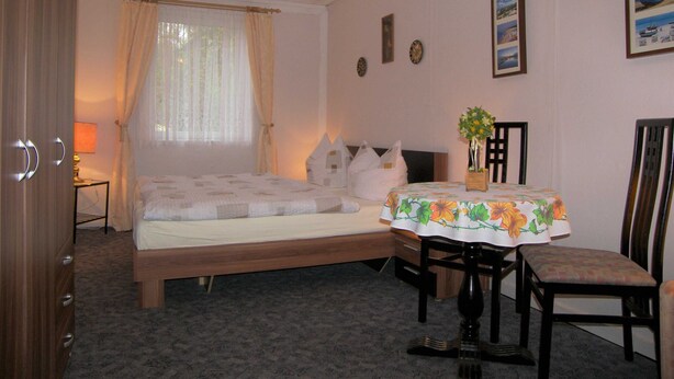 A bedroom with a bed, a bedside table, a chair, a small table with a floral tablecloth, and a window with curtains. - Bungalow Lange with 2 bedrooms - Bungalow Lange (Heringsdorf)
