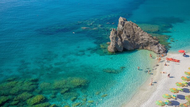 Ein Strand mit klarem türkisblauem Wasser, Felsformationen und einem Sandufer mit Sonnenschirmen und Liegestühlen.
