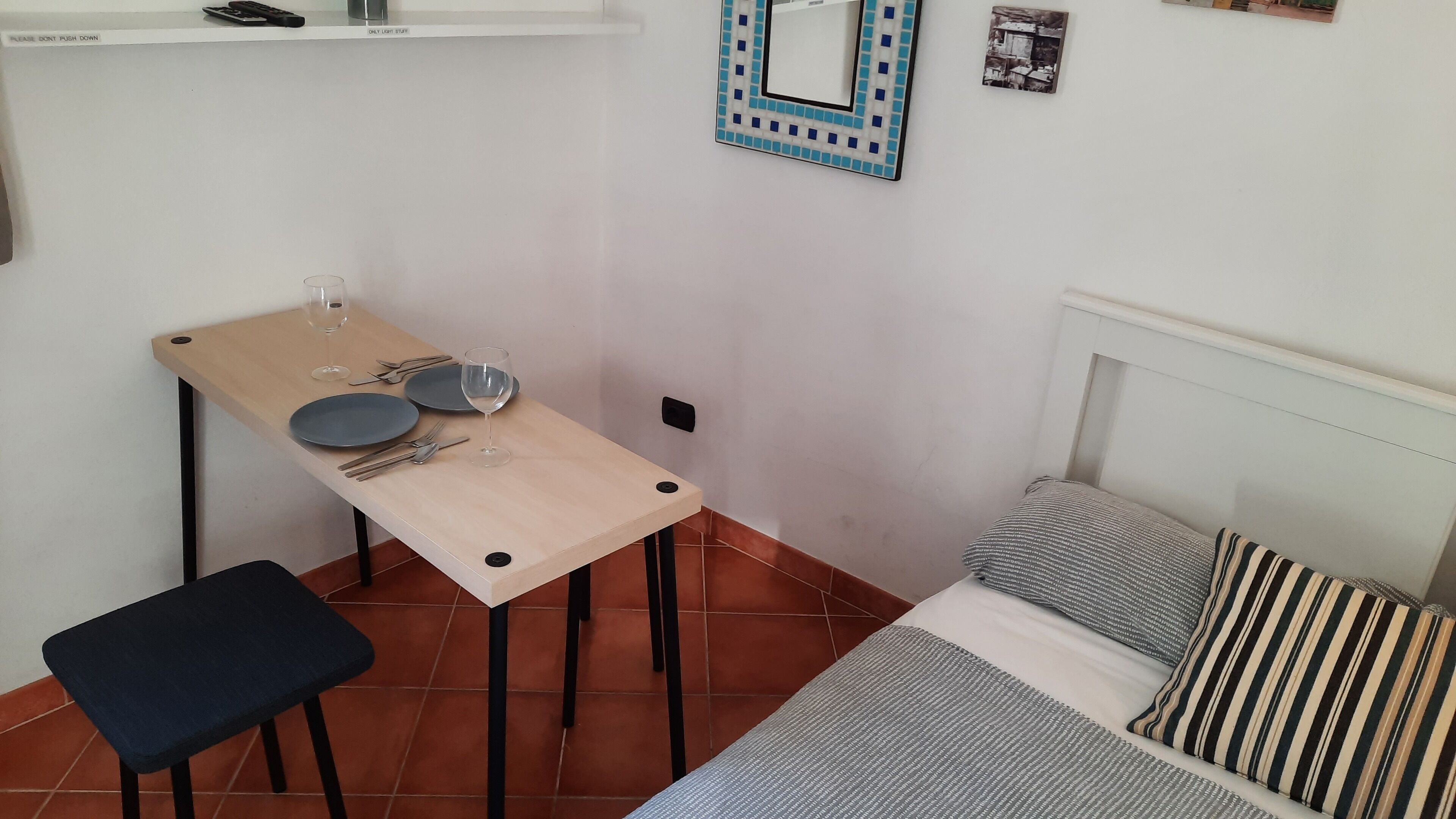 A small, neatly arranged room with a bed, a table with plates, cutlery, and a wine glass, a chair, and a mirror on the wall.