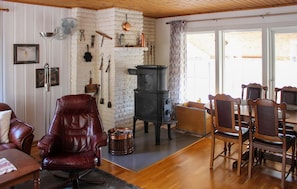 Un salon chaleureux avec un fauteuil en cuir, une table à manger en bois entourée de chaises, un poêle noir et divers ustensiles de cuisine accrochés au mur.