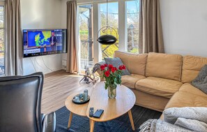 Un salon comprenant un canapé, une table basse ornée d’un vase de fleurs, un téléviseur affichant les prévisions météorologiques et une vue sur l’extérieur grâce à de grandes fenêtres.