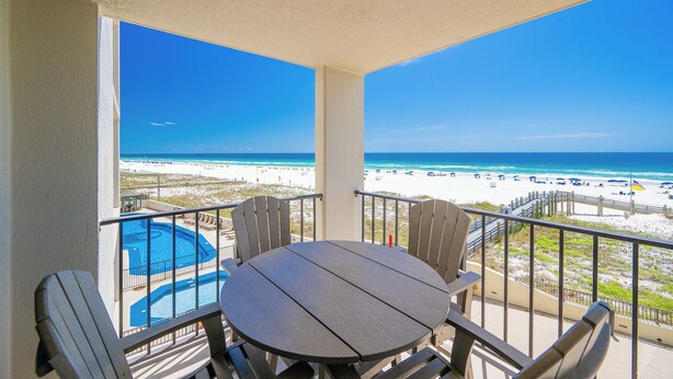 A balcony with a view of the beach, a pool, and outdoor seating.