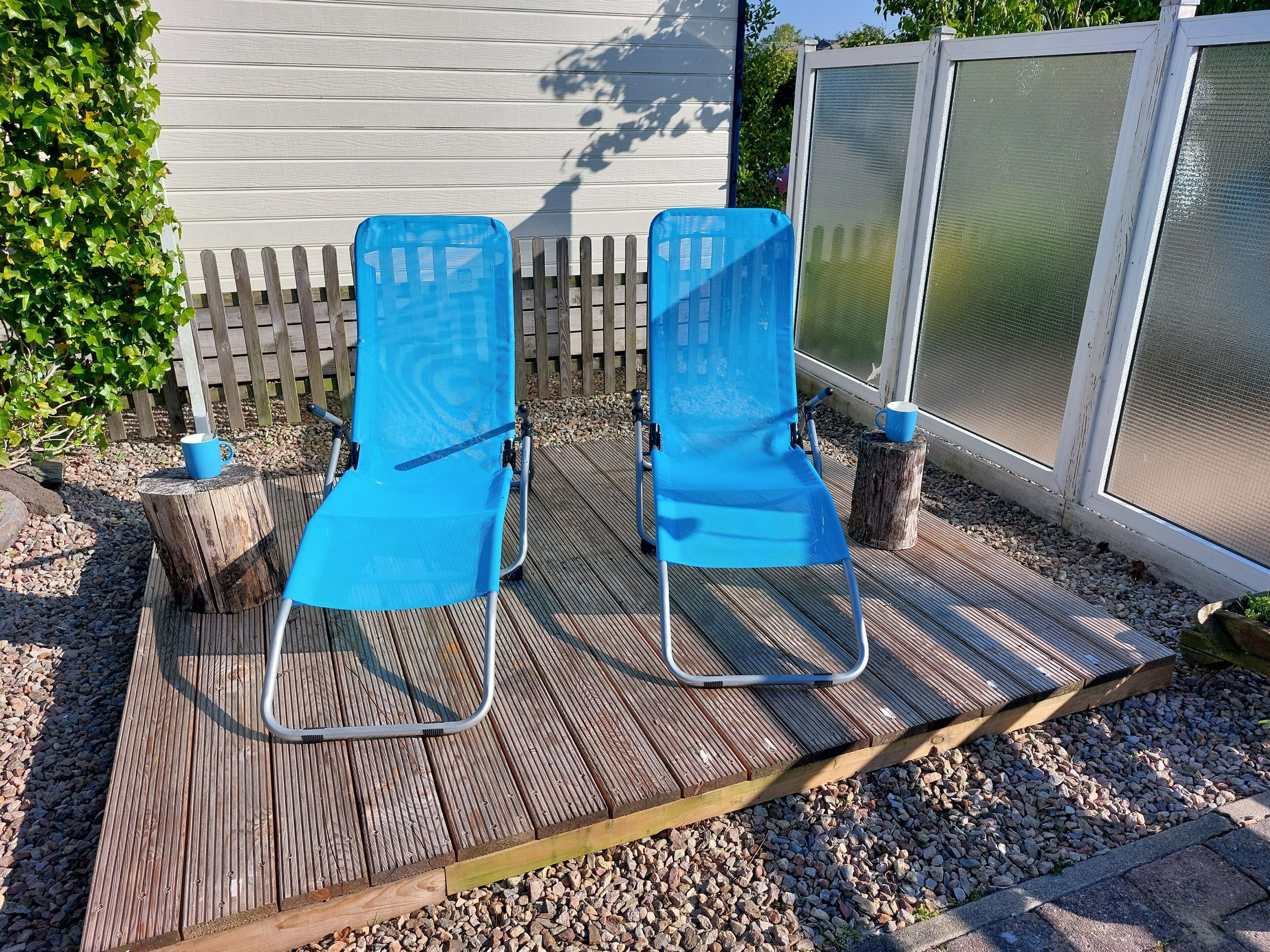 Two blue lounge chairs on a wooden deck with a white fence and greenery in the background.