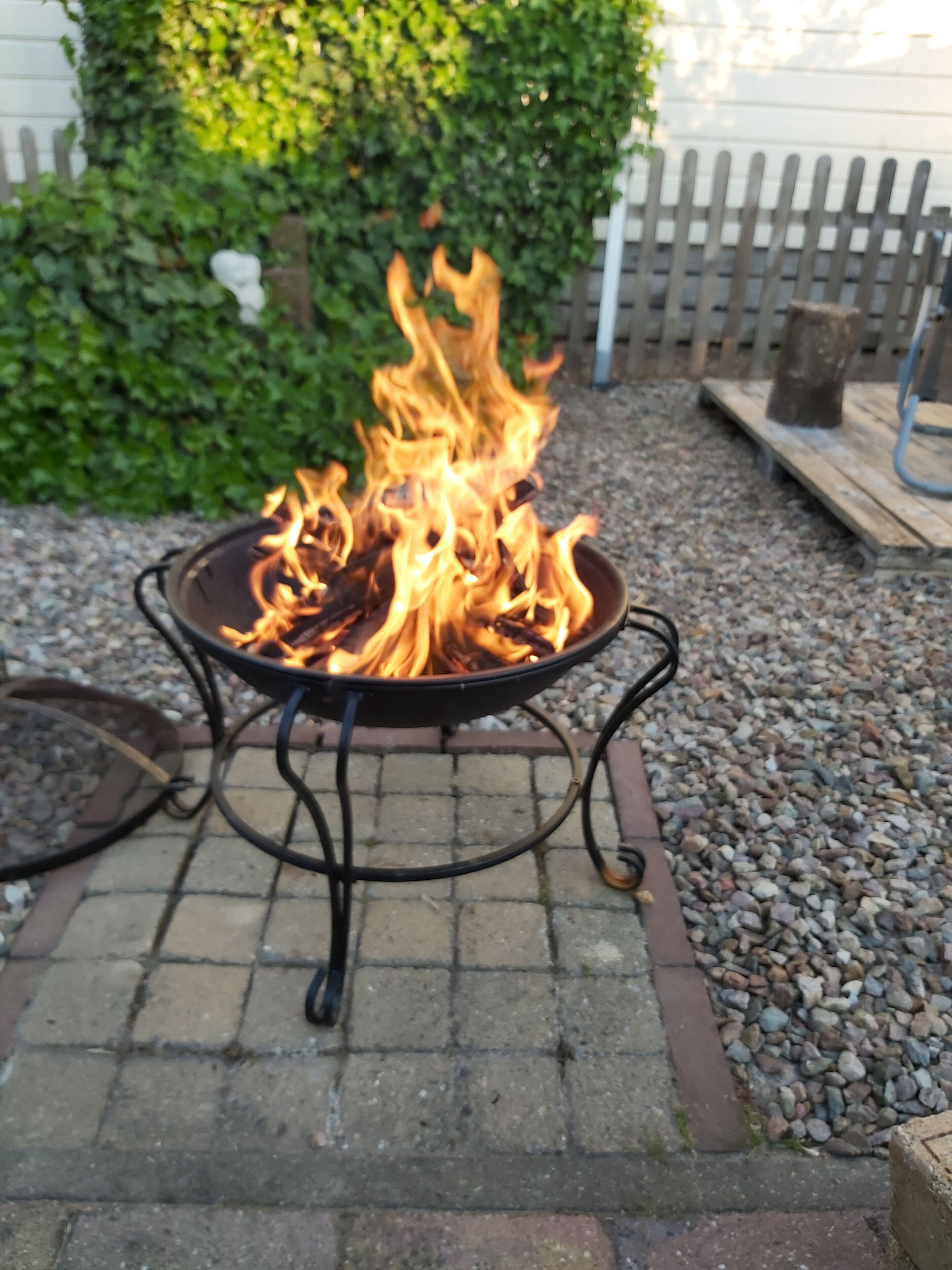 Un foyer métallique avec des flammes, entouré d’une terrasse pavée avec du gravier et une clôture en bois en arrière-plan.