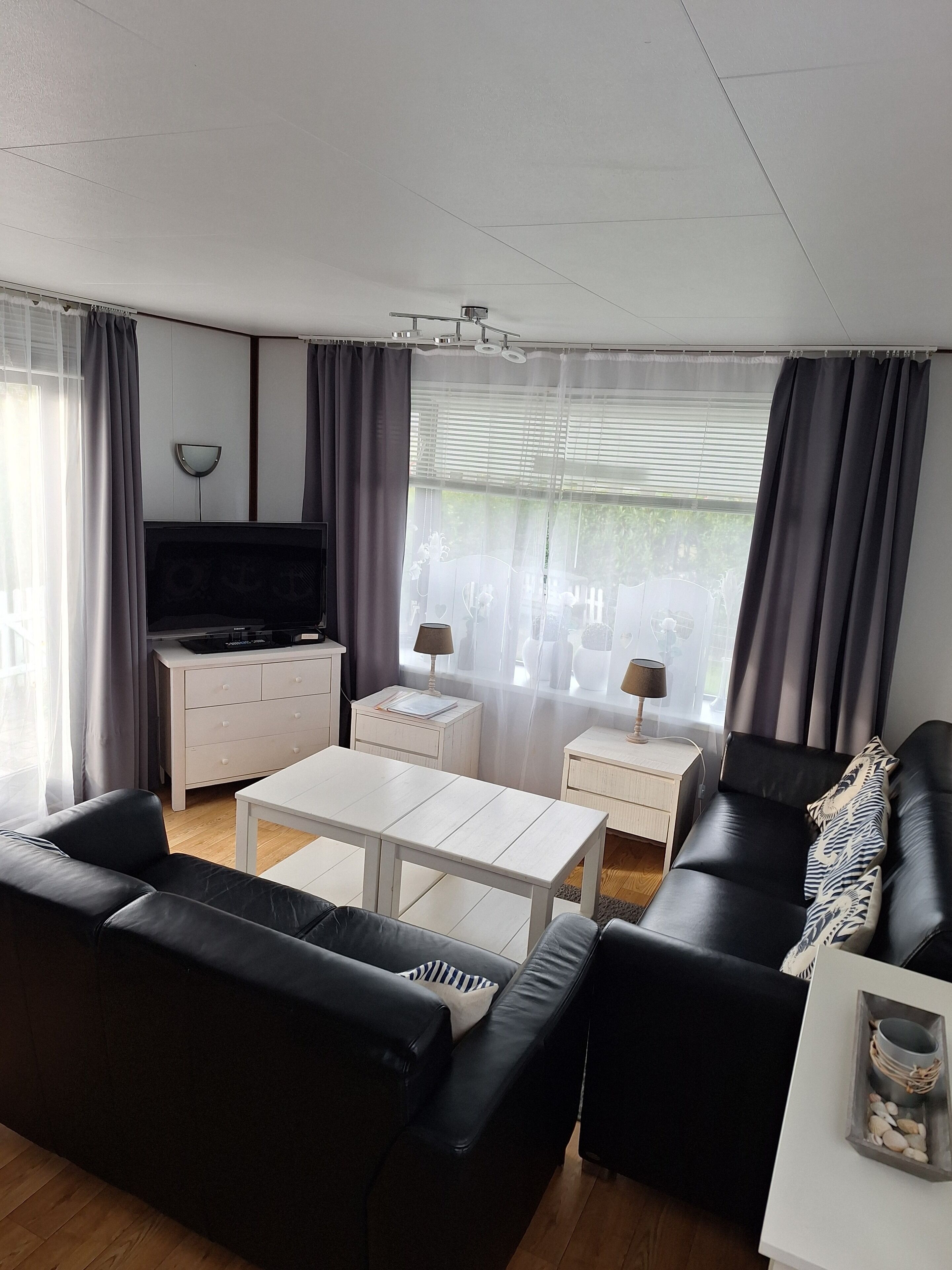 A modern living room with a flat-screen TV, a white coffee table, black leather sofas, and a white side table with a lamp.