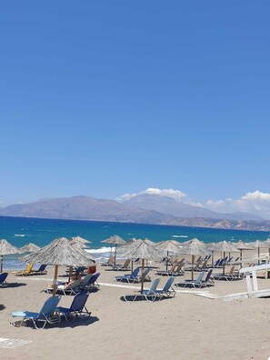 A beach with rows of sun loungers and thatched umbrellas, a clear blue sky, and mountains in the background.