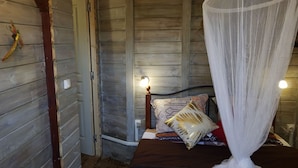 Une pièce en bois avec un lit, une table de chevet, une lampe fixée au mur et une moustiquaire.