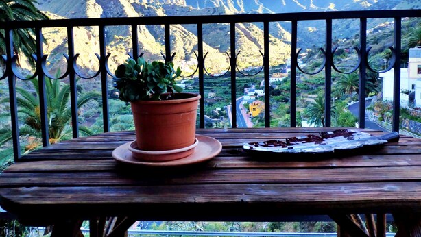 Ein Holztisch mit einer Topfpflanze und einem Teller Essen auf einem Balkon mit Blick auf die Berge.