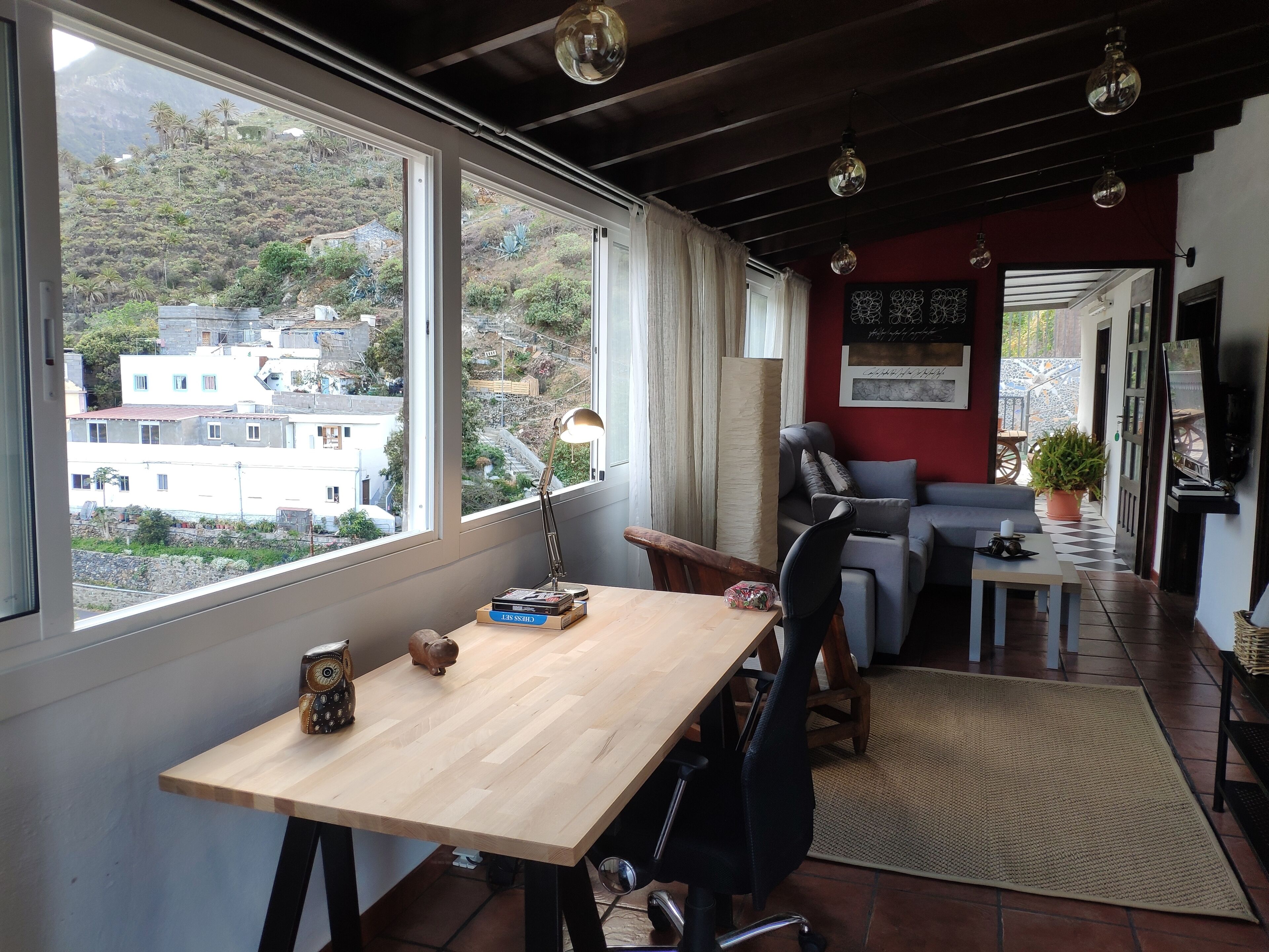 A cozy room with a wooden desk, a comfortable sofa, and a large window offering a view of a hillside.