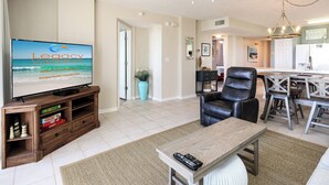 A living room with a television, a wooden entertainment unit, a black leather chair, a wooden coffee table, and a kitchen area in the background. - Island Princess 204-Updated Beachfront with FAST Internet-Free Beach Service (Fort Walton Beach)