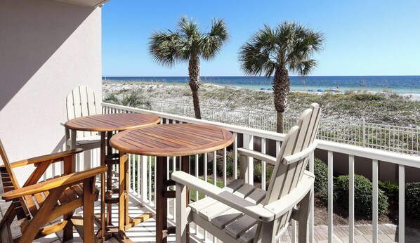 Un balcon avec deux tables en bois et des chaises blanches, donnant sur une plage bordée de palmiers et surplombant un ciel dégagé.