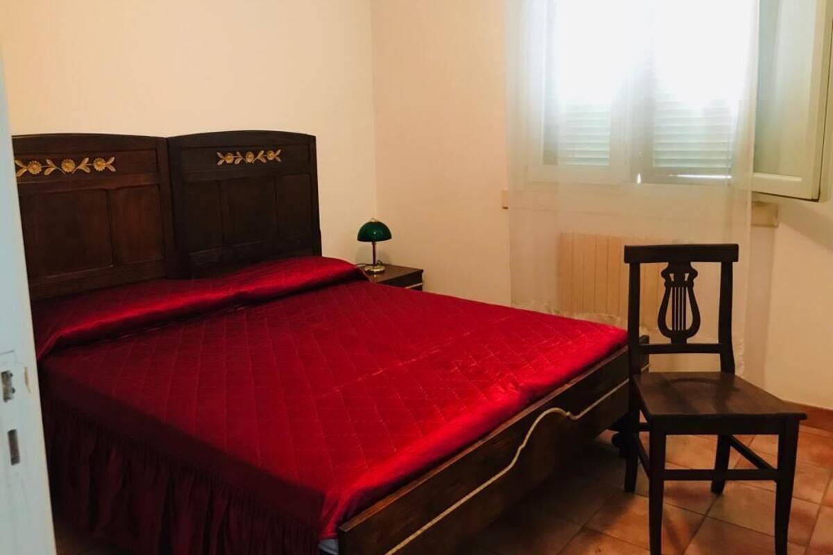 A bedroom with a wooden bed, a red bedspread, a nightstand with a lamp, a chair, and a window with blinds.