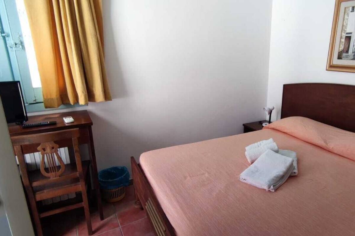 A bedroom with a bed, a wooden chair, a desk with a computer, and a window with curtains.