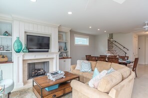 A living room with a fireplace, a television, a sofa, and a dining area with chairs and a table. - 120 Luxury Sandcastle Home w/ Pool, Elevator & Bunks (Hilton Head Island)