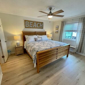 A bedroom with a wooden bed, a ceiling fan, a beach-themed sign, and a window with curtains. - Duneside I 104 - Oceanfront Complex (Oceanview Unit) in Garden City Beach (Garden City)
