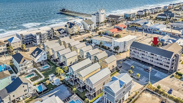A coastal residential area with a pier extending into the ocean, featuring a mix of single and multi-story houses, a parking lot, and a swimming pool area.