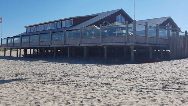 Un bâtiment situé en bord de mer, doté d’une terrasse en bois et de grandes fenêtres, qui ressemble probablement à un hôtel ou à un complexe touristique.