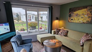 Un salon comprenant un canapé, un fauteuil et un pouf, une télévision et une grande fenêtre donnant sur une maison et un jardin.