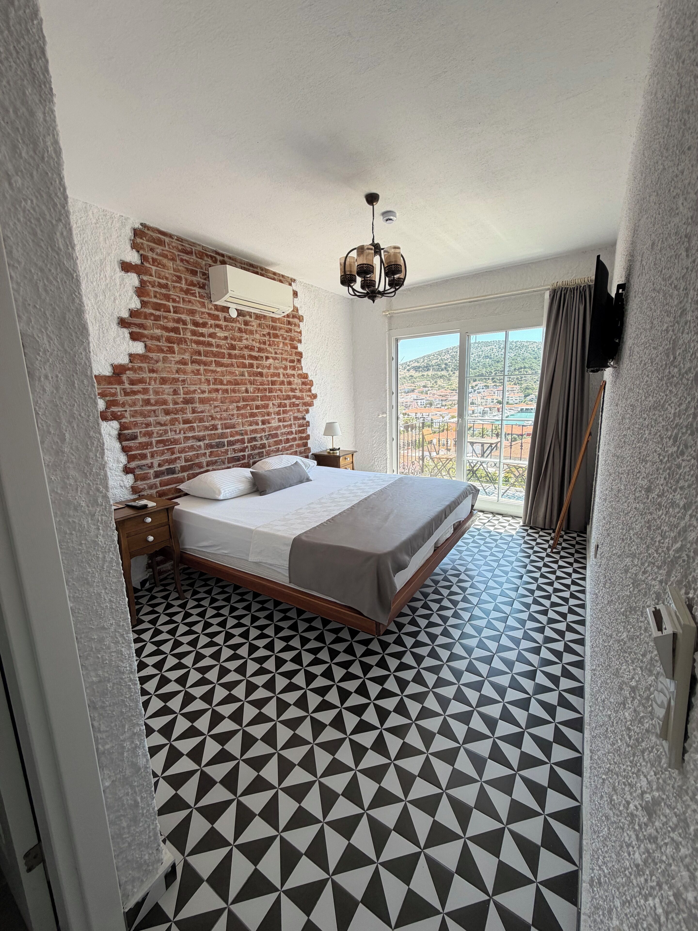 Ein Schlafzimmer mit Backsteinwand, einem Bett mit weißer und grauer Bettdecke, einer Kristallleuchtre und einem geometrisch gemusterten Boden.