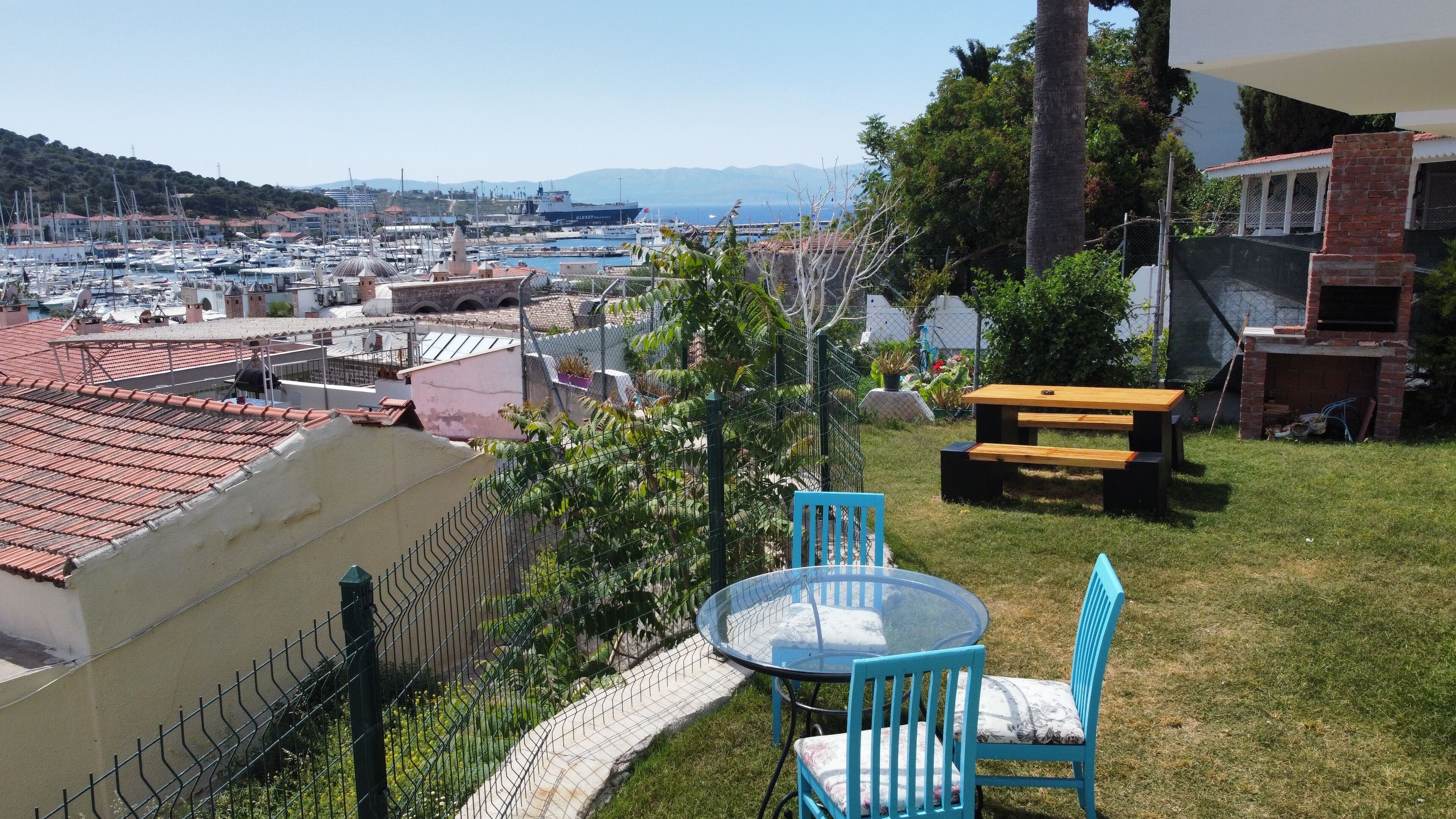 A patio with a glass table and blue chairs, overlooking a marina with boats and a distant cityscape.