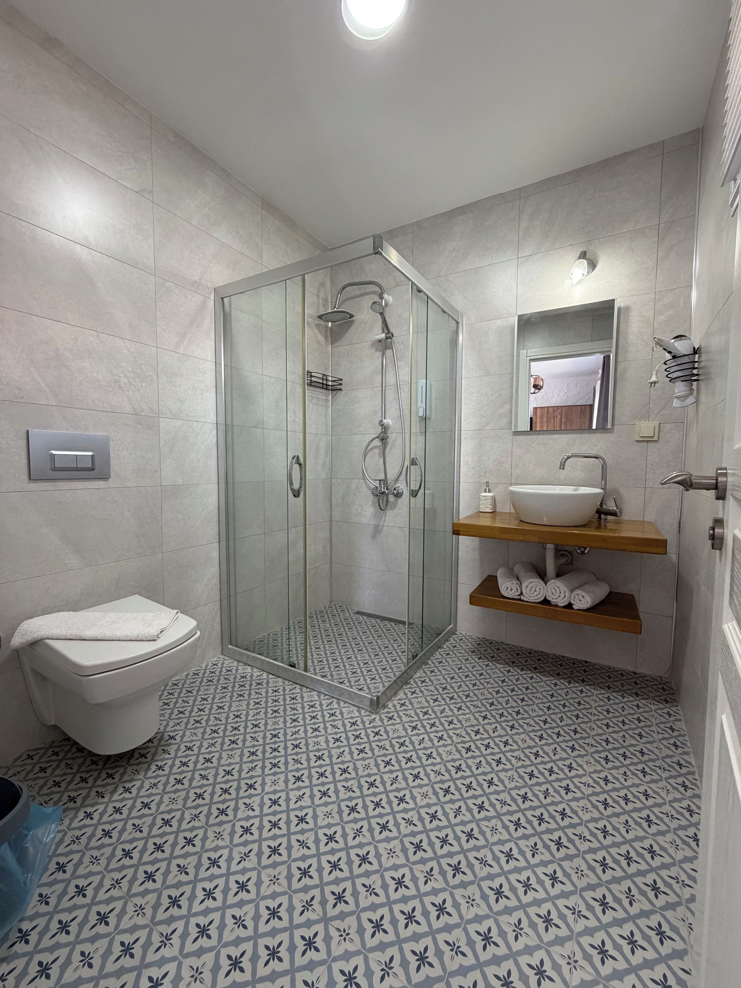 A modern bathroom with a glass-enclosed shower, a toilet, a sink with a mirror, and patterned floor tiles.