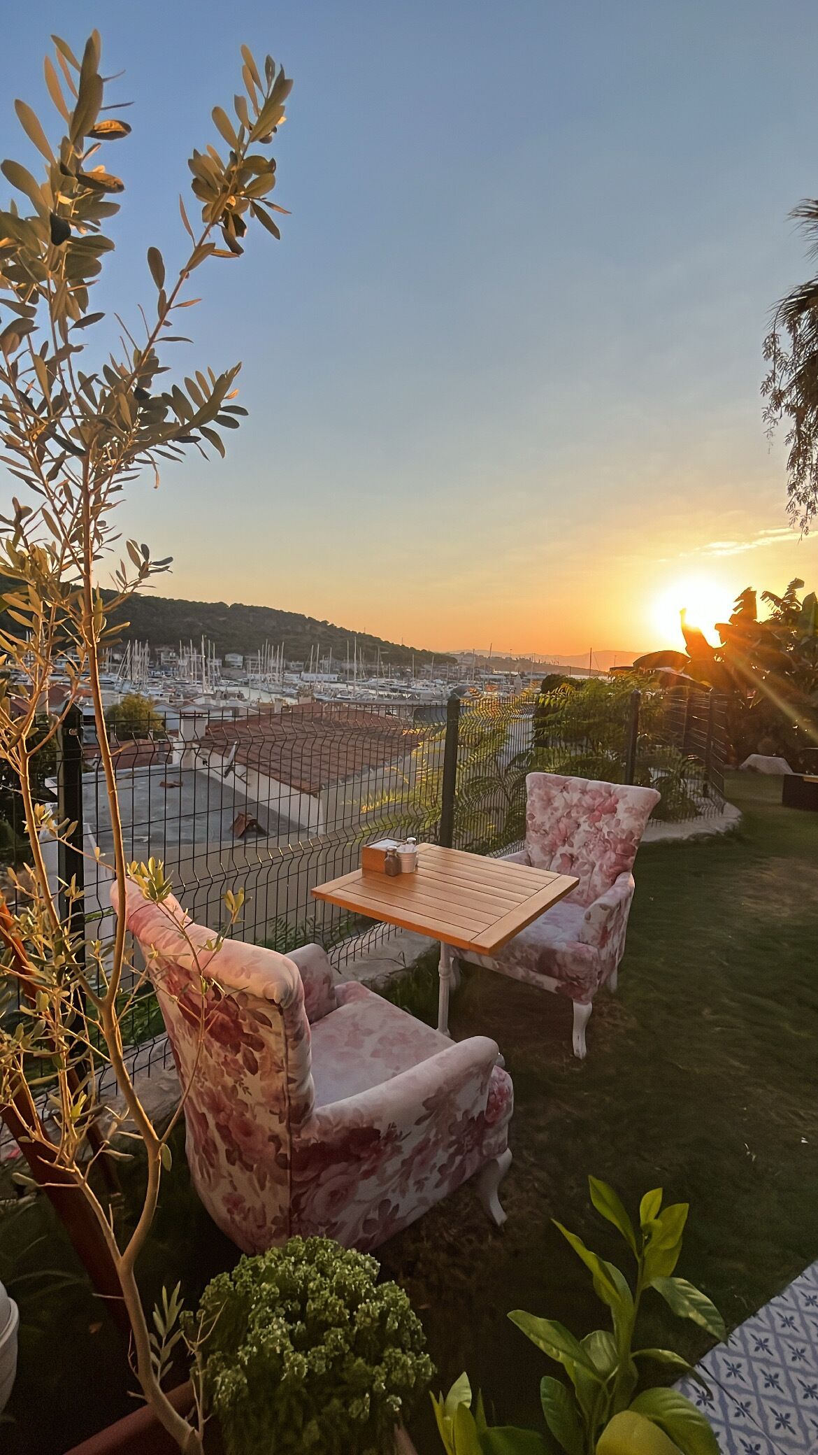 Um pátio com duas cadeiras florais e uma mesa pequena, com vista para uma marina com barcos e um pôr do sol ao fundo.