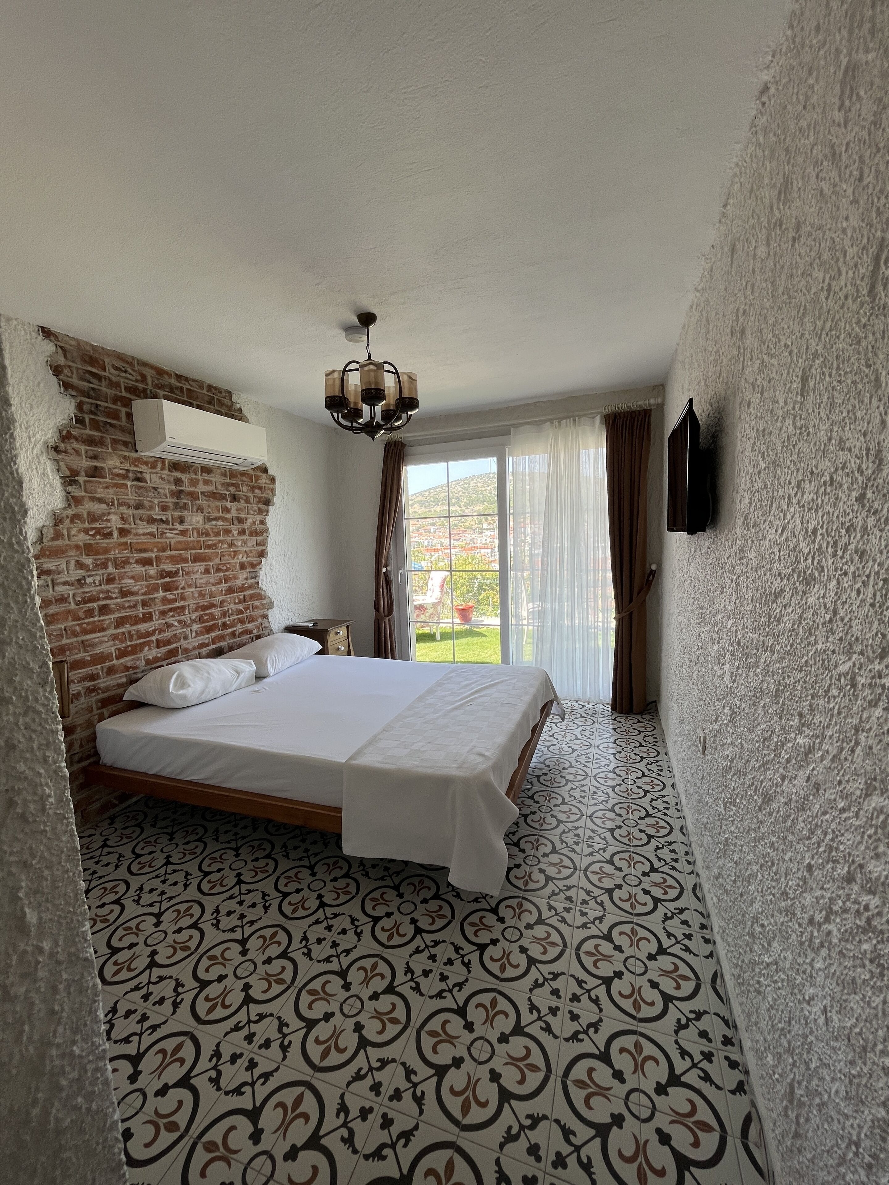 Un dormitorio con pared de ladrillo, una cama con cubrecama blanco, una araña, un televisor montado en la pared y un piso con diseño.