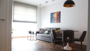 Una sala moderna con un sofá gris, una mesa de comedor con sillas, un ventanal con persianas y un cuadro abstracto y colorido en la pared.