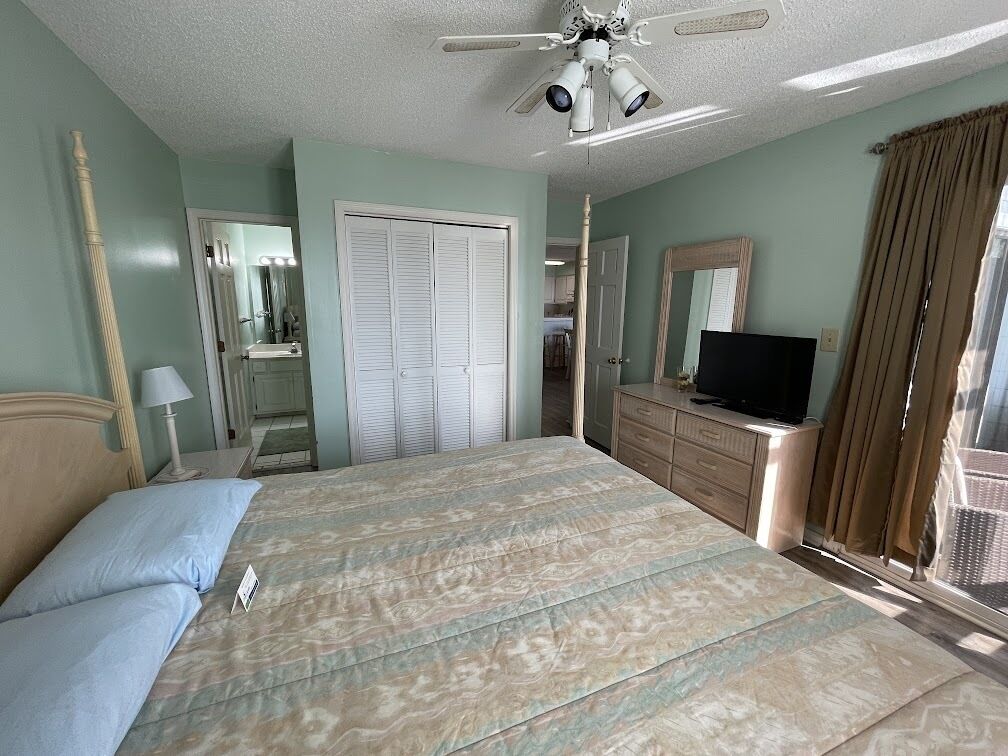 Une chambre à coucher comprenant un lit, une commode, un téléviseur, un ventilateur de plafond et une fenêtre avec des rideaux.