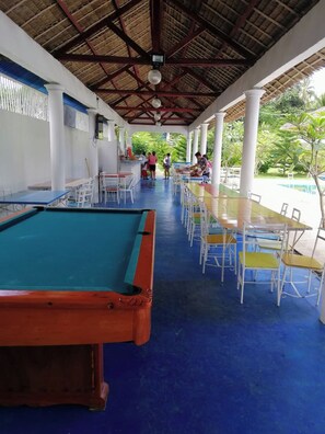 A pool area with a pool table, multiple tables, and chairs, surrounded by a covered walkway with a thatched roof. - Sweet Water Resort (Zamboanguita)