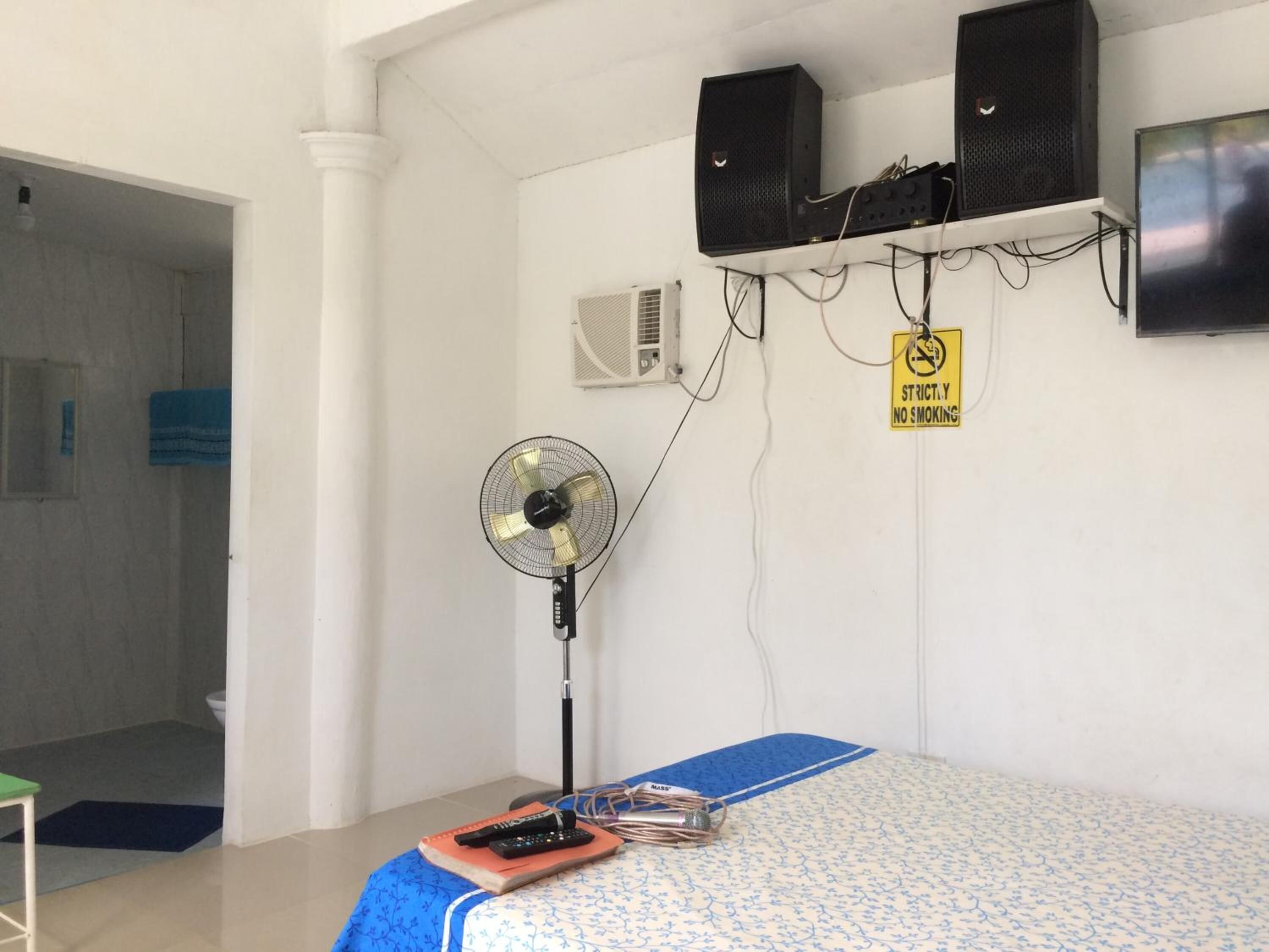 Une chambre avec un lit, un ventilateur sur pied, deux haut-parleurs, une télévision et un panneau « interdiction de fumer » accroché au mur.