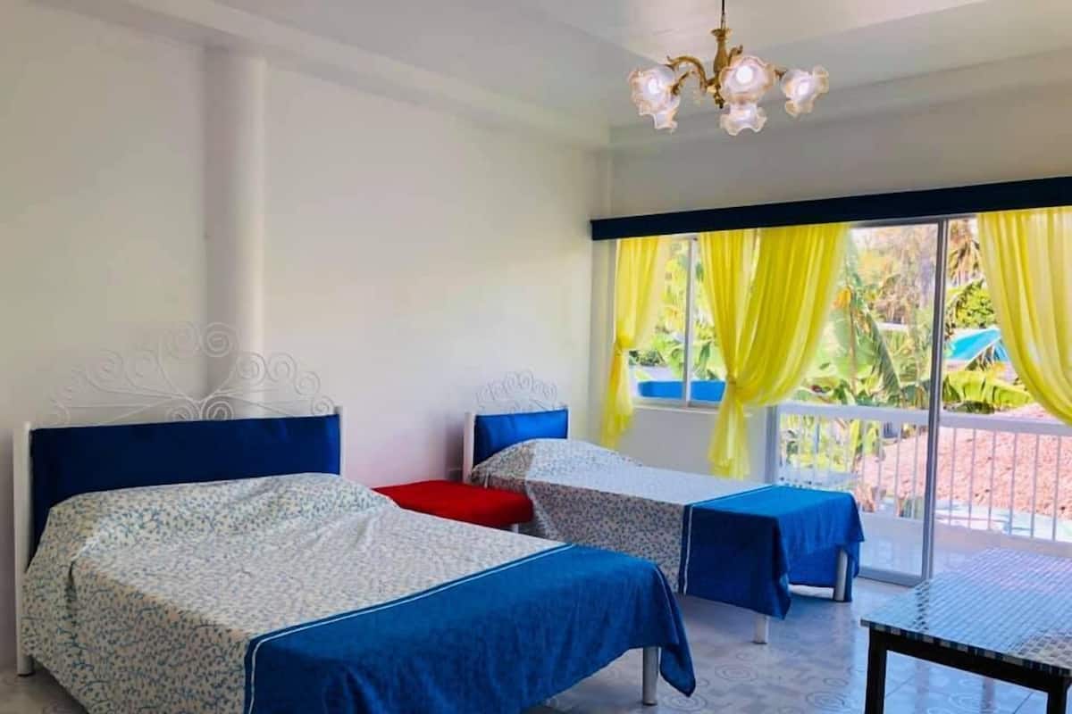 A room with two beds, a chandelier, and a view of greenery through a window with yellow curtains.